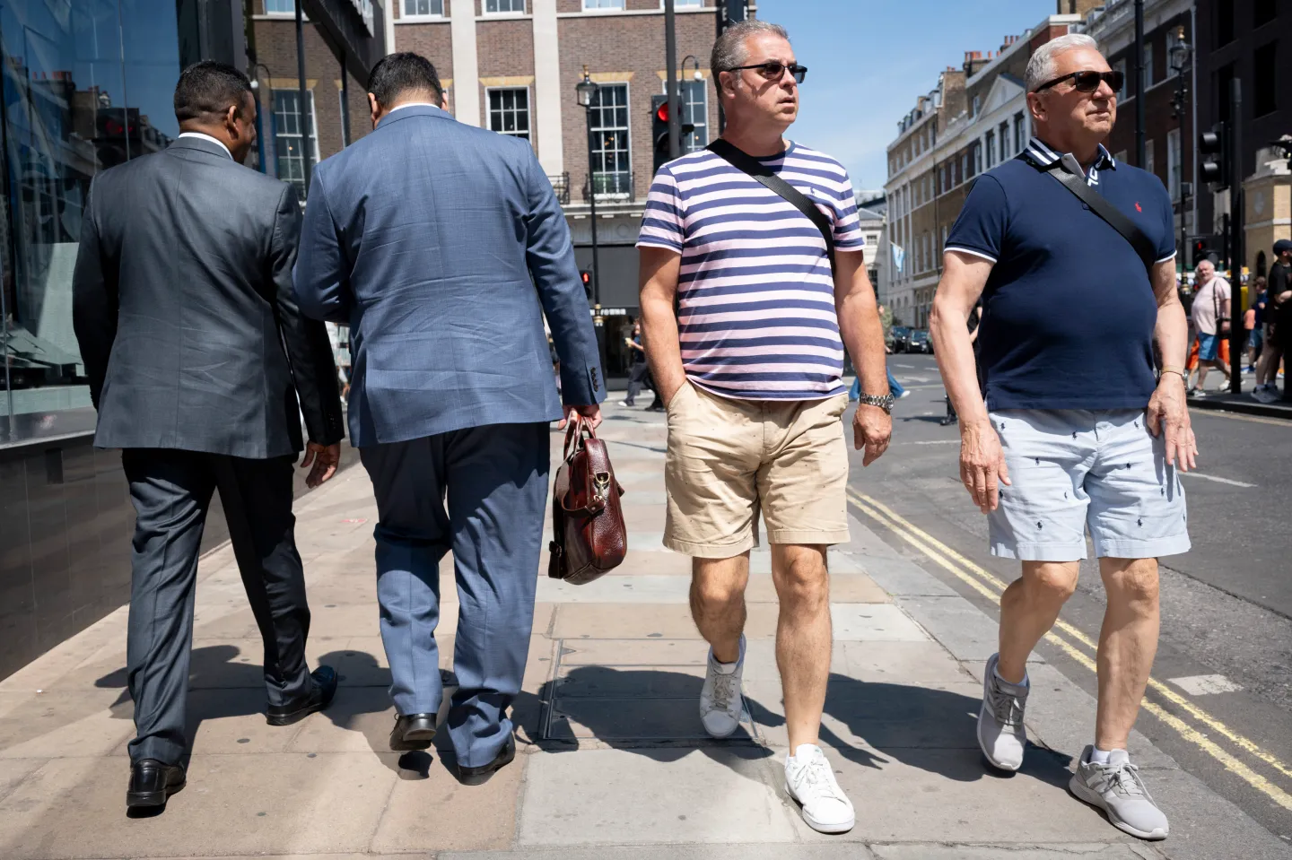 From the rear we see two men wearing impractical business suits walking past two others wearing cooler, more sensible shorts and casual wear for the summer heatwave, on 15th June 2023, in London, England. (Photo by Richard Baker / In Pictures via Getty Images)